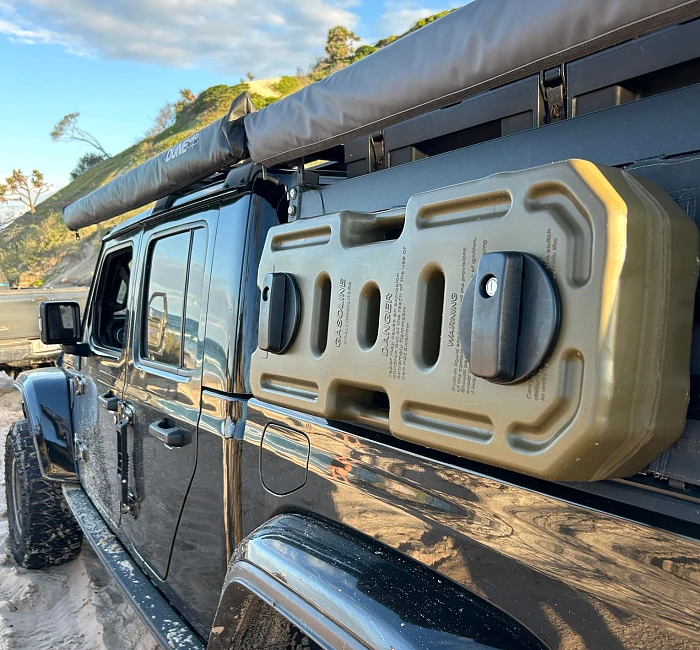 A heavy-duty jerry can mounted to the roof rack of a Jeep Gladiator fitted with an ARB awning, parked at the beach